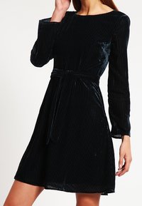 Robe en velours noir à manches longues, texture côtelée horizontale, taille cintrée et ceinture assortie, s'évasant doucement au niveau de l'ourlet.