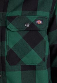 Chemise à carreaux verte et noire à boutons, avec une poche à rabat, présentant une fermeture à pression et une petite étiquette de marque sur la poitrine.