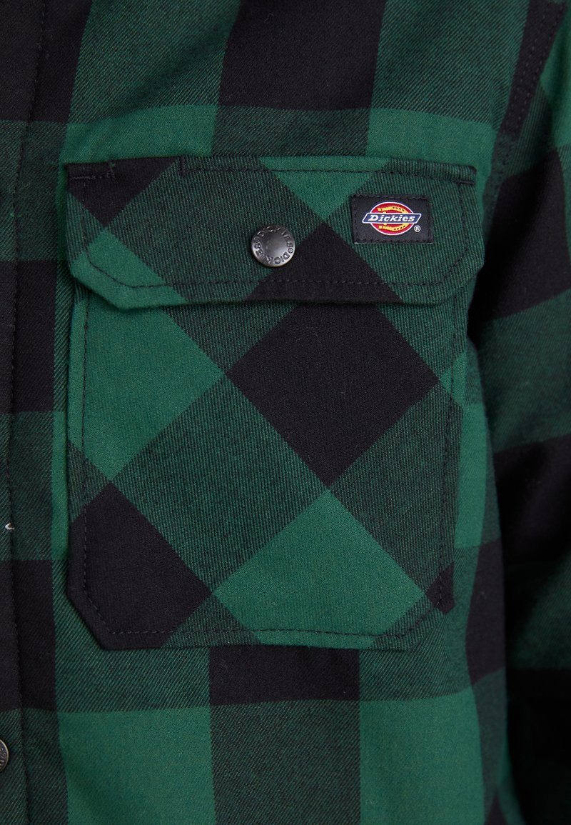 Camicia a quadri verde e nera con taschino a patta, dotata di chiusura a scatto e un piccolo etichetta del brand sul petto.