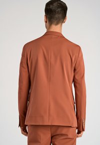 Blazer color óxido, corte entallado, tejido suave, abertura trasera, puños de cinco botones. La vista trasera resalta una silueta ordenada y líneas limpias.