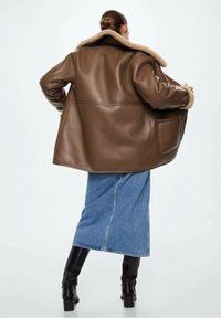 Giacca di pelle marrone con colletto in pelliccia sintetica, vestibilità ampia e tasche laterali. Abbinata a una gonna di jeans e stivali neri alti fino al ginocchio.