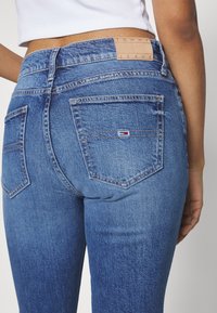 Vaqueros de mezclilla azul con un diseño ajustado, que presentan un parche de cuero color beige etiquetado "TOMMY JEANS" y un pequeño logo en el bolsillo trasero.