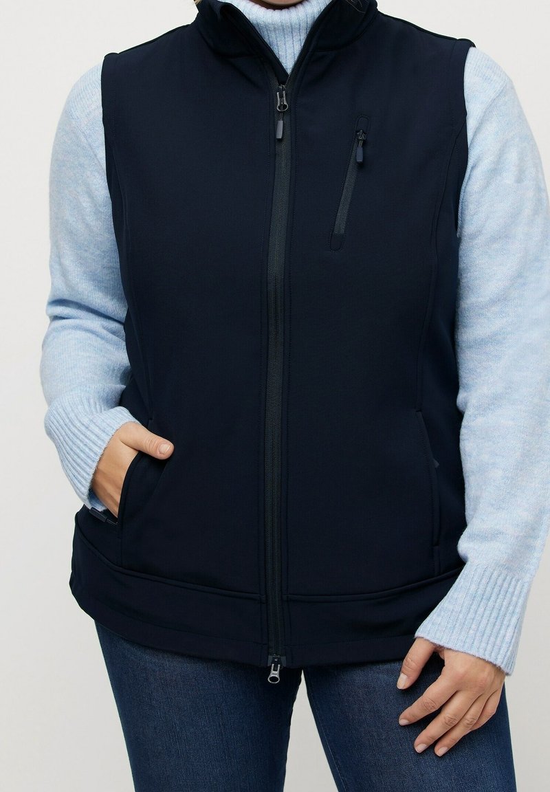 Marineblauer ärmelloser Weste mit hohem Kragen, Frontreißverschluss und zwei Seitentaschen; über einem hellblauen Pullover getragen; glatte Textur.