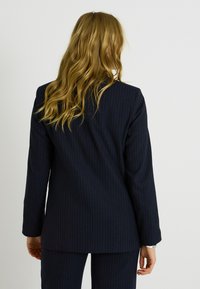 Blazer de rayas marineras con mangas largas, corte recto y textura suave. Presenta un cuello sencillo y un ligero acolchado en los hombros.