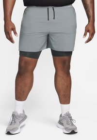 Homme portant un short de sport gris avec un short de compression noir en dessous, des chaussettes blanches montantes et des chaussures de course grises sur fond blanc.
