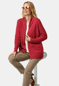 Roter Cardigan mit offener Vorderseite, gerippten Bündchen und zwei Taschen, kombiniert mit beigen Hosen und braunen Absätzen. Das Model trägt Sonnenbrillen und lächelt.