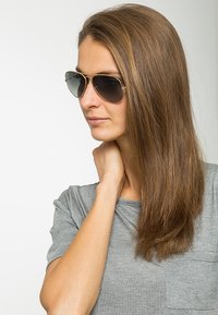 Occhiali da sole con lenti scure e montatura in sottile metallo dorato. Capelli lunghi castani, indossando una maglietta grigia con taschino. Design semplice e moderno.