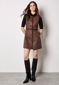 Brązowa zamszowa mini sukienka z pionowymi przeszyciami i wyrazistą sylwetką, zestawiona z czarnymi skórzanymi butami do kolan.