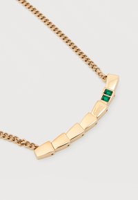 Χρυσό κολιέ αλυσίδα με έξι τμήματα τραπεζοειδούς σχήματος, δύο από τα οποία διαθέτουν πράσινες ορθογώνιες πολύτιμες πέτρες, τοποθετημένα σε καμπυλωτό μοτίβο σε ανοιχτό φόντο.