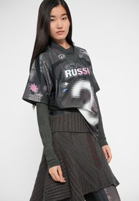 Mlada žena s dugom tamnom kosom koja nosi crnu grafičku oversized majicu preko teksturirane tamne pletene suknje i dugih rukava.