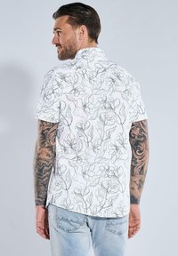 Kurzarmhemd aus weißem Stoff mit schwarzer floraler Linienkunst. Bemerkenswerte Details sind ein Kragen und seitliche Schlitze am Saum.