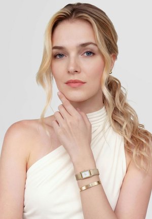 Femme blonde aux cheveux ondulés portant un haut blanc asymétrique, avec une montre-bracelet dorée et un bracelet, touchant légèrement son menton avec ses doigts.