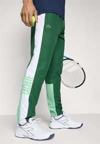 Pantaloni de trening verzi și verde deschis cu panouri laterale albe, prezentând imprimeul "LACOSTE" și un logo, asortați cu pantofi sport albi.