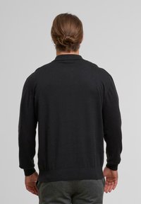 Mann mit hellbraunem Haar, das zu einem Dutt gebunden ist, trägt einen schwarzen Pullover und graue Hosen und steht mit dem Rücken zu einer einfarbigen Wand.