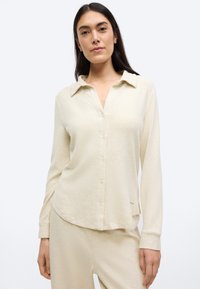 Krém színű, gombos ing, galléros nyakkivágással, hosszú ujjal és puha tapintással. Hasonlóan puha nadrággal párosítva.