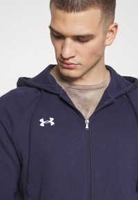 Marineblauwe rits-hoodie van zacht materiaal met een capuchon, voorzien van een wit Under Armour-logo op de linkerborst en een rits aan de voorkant.