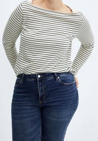 Personne portant une chemise blanche à manches longues avec des rayures horizontales noires et un jean bleu foncé, debout les mains dans le dos.