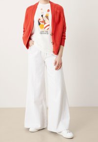 Röd ribbad kofta med träknappar, vit grafisk t-shirt med färgglad handdesign och vida vita byxor, kombinerat med vita sneakers.