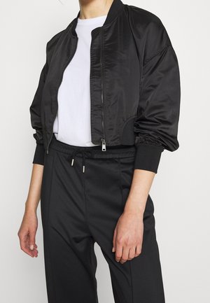 Personne portant une veste bomber noire courte sur une chemise blanche et un pantalon noir taille haute avec une taille à cordon.