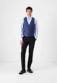Colete de lã azul com cinco botões, usado sobre uma camisa azul clara. Calças pretas e sapatos pretos completam o conjunto. Design moderno e simples.