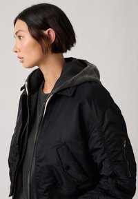 Νεαρός με κοντά σκούρα μαλλιά, που φορεί μαύρο στρωματοποιημένο μπουφάν με φερμουάρ και σκούρα κουκούλα, στραμμένος προς τα αριστερά μπροστά σε ένα απλό φόντο.