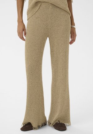 Pantalones anchos de punto color beige con dobladillos desgastados, usados con zapatos planos marrón oscuro en una persona de pie.