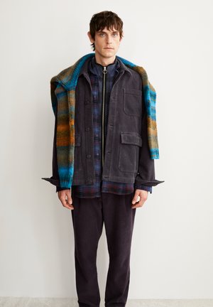 Giacca di velluto a coste grigia scura con due tasche frontali, indossata sopra una camicia a quadri blu e rossa, completata da un colorato scialle di maglia.