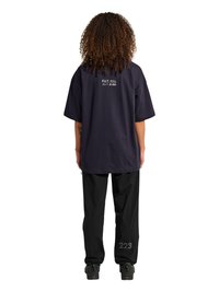 Osoba o kręconych włosach nosi luźny ciemny t-shirt z małym napisem z tyłu oraz czarne spodnie z numerem „223” na łydce, stojąc na białym tle.