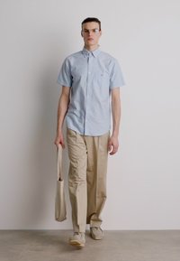 Camisa azul-claro de botão de mangas curtas, calças largas bege e ténis bege. Modelo segura uma bolsa de lona, em pé sobre um fundo neutro.