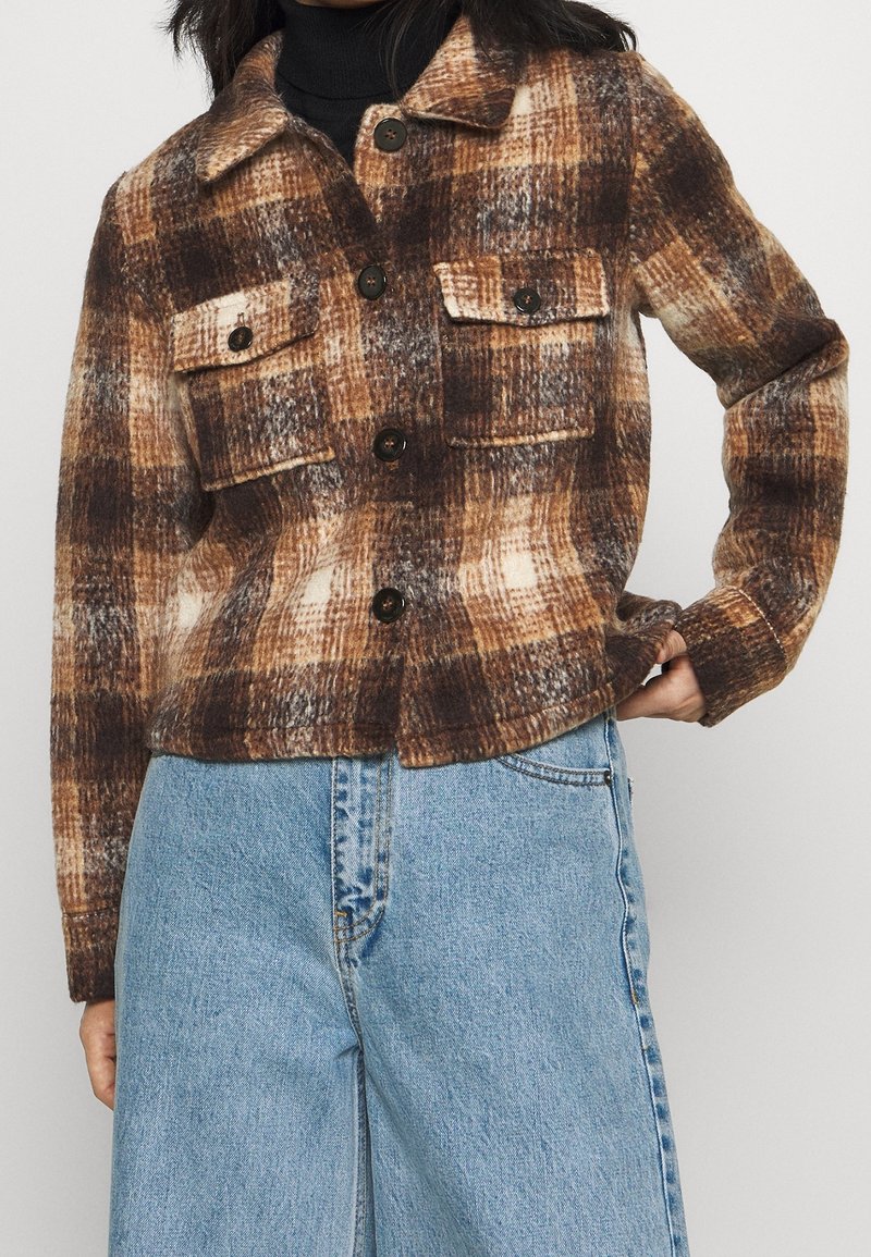 Manteau en flanelle à carreaux marron et crème avec un col, fermeture à boutons sur le devant, et deux poches poitrine. Tissu texturé avec un motif bien visible.