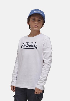 Garçon portant un shirt à manches longues blanc de Von Dutch, une casquette bleue Von Dutch et un pantalon cargo noir, debout devant un fond uni.