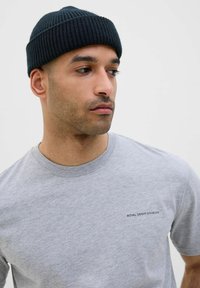 Grey cotton t-shirt with crew neck and short sleeves; features small black logo text on the left chest. Worn with a dark ribbed beanie.