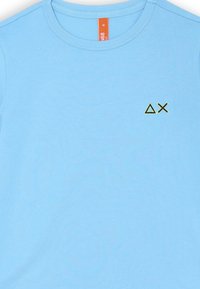 T-shirt di cotone azzurro chiaro con colletto a girocollo e un logo geometrico ricamato verde sul lato sinistro del petto. Texture liscia.