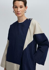 Marineblauwe en beige oversized top met een textuurafwerking. Heeft een ronde halslijn, lange mouwen en knoopaccenten op één schouder.