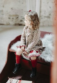 Strickkleid mit roten Herzmustern, beiger Grundfarbe, Rüschensaumkragen, kombiniert mit roten kniehohen Socken und glänzenden schwarzen Stiefeln. Weiches Kunstfell-Sitzpolster.