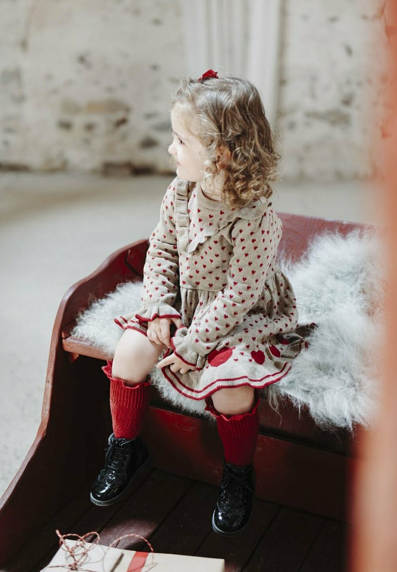 Strickkleid mit roten Herzmustern, beiger Grundfarbe, Rüschensaumkragen, kombiniert mit roten kniehohen Socken und glänzenden schwarzen Stiefeln. Weiches Kunstfell-Sitzpolster.