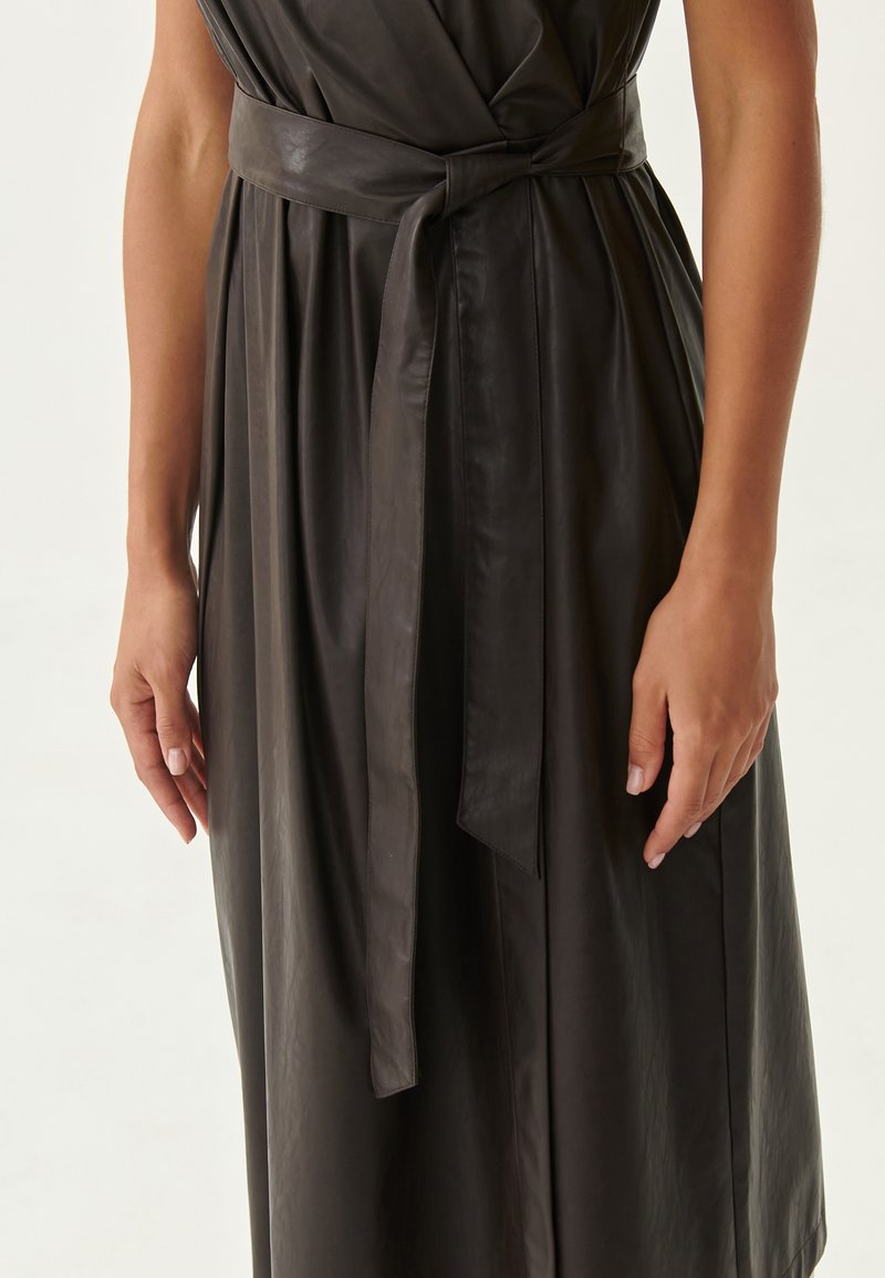 Abito in pelle nera con design a portafoglio, dotato di una cintura in vita e una texture liscia. Sono visibili maniche lunghe e dettagli di tessuto fluido.