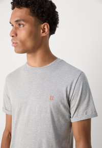 Camiseta gris de manga corta hecha de algodón, con un pequeño "II" bordado en color naranja en el lado izquierdo del pecho y un cuello redondo.