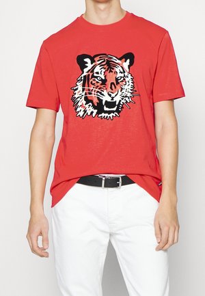 Mężczyzna w czerwonym t-shircie z krótkim rękawem ozdobionym grafiką ryczącego tygrysa, białe spodnie i czarny pasek na jednolitym tle.