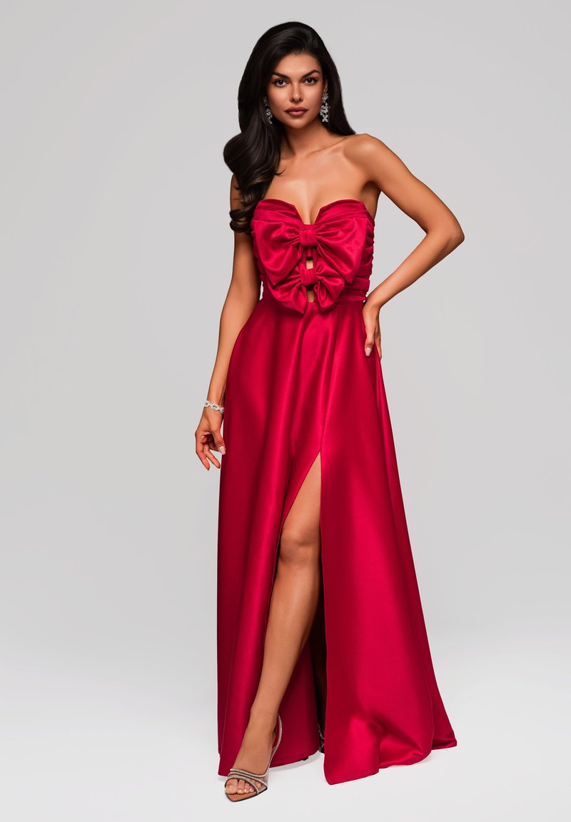 Robe en satin rouge sans bretelles avec un grand nœud à la poitrine, présentant une fente haute sur la cuisse et une silhouette lisse et fluide. Accentuée avec des sandales.