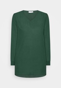 Grön blus med långa ärmar och V-ringning, tillverkad av ett slätt tyg. Rundad nederkant och knappade manschetter med minimalistisk design.