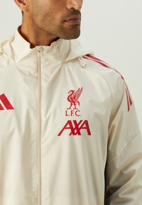 Cremefarvet letvægtsjakke med hætte, der har rød Liverpool FC-logo, AXA-logo og røde Adidas-striber på skuldrene.