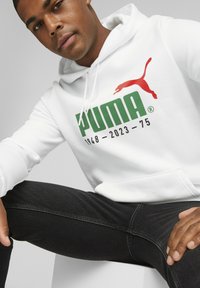 Sudadera blanca con un logo de PUMA verde y acentos rojos, y jeans negros. La sudadera tiene un bolsillo tipo canguro y capucha con cordón.