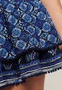 Tejido azul con estampado en diferentes tonalidades de azul, con un dobladillo volado y un borde con pompones. Etiqueta visible con el texto "Superdry."