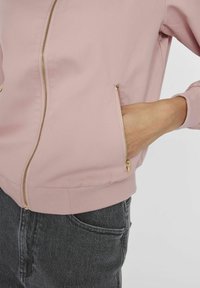 Personne portant une veste zippée rose clair avec des zippers dorés et un jean gris foncé, main placée dans la poche de la veste sur fond blanc.