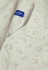 Bijela košulja s dugim rukavima s složenim cvjetnim vezom i izrezanim uzorcima. Ima ovratnik i oznaku s natpisom "Jack & Jones" veličina L.