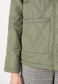 Fållad grön jacka med en fyrkantig bröstficka, med ett diamantmönster och trekvartslånga ärmar, tillsammans med rutiga byxor.