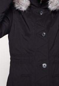 Svart isolerad jacka med huva fodrad med fake päls, knappstängning och två framfickor med texturerad sömnadsdetalj.