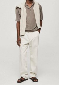 Homme portant un polo beige, un pull en tricot crème posé sur les épaules, un pantalon blanc et des sandales noires ouvertes, debout avec une main dans la poche.