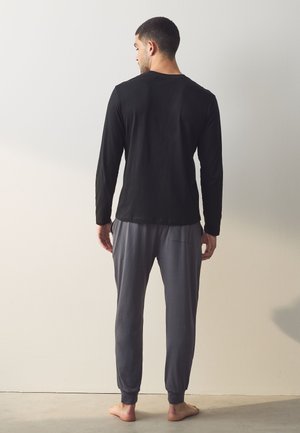 Homme debout pieds nus, face à un mur blanc, portant une chemise noire à manches longues et un pantalon de jogging gris.